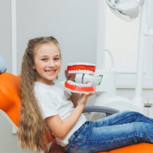 Little girl sitting on a dental chair holding a model of jaws in a dental clinic.