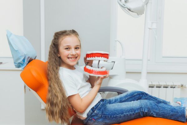 Little girl sitting on a dental chair holding a model of jaws in a dental clinic.